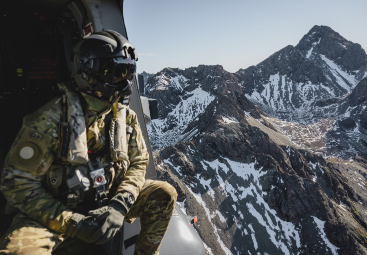 A helicopter loadmaster in full Air Force uniform including a flight helmet looks out the side of the aircraft to snow-capped mountains.