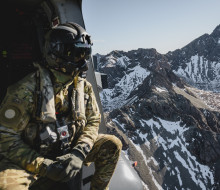 A helicopter loadmaster in full Air Force uniform including a flight helmet looks out the side of the aircraft to snow-capped mountains.