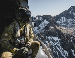A helicopter loadmaster in full Air Force uniform including a flight helmet looks out the side of the aircraft to snow-capped mountains.