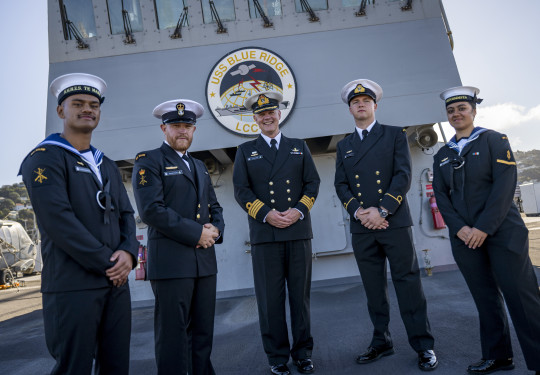 Five Navy sailors stand in uniform in front of a large "USS Blue Ridge". The ship casts a shadow on the sailors on a cold sunny day.