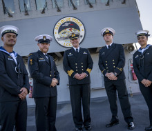 Five Navy sailors stand in uniform in front of a large "USS Blue Ridge". The ship casts a shadow on the sailors on a cold sunny day.