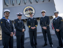 Five Navy sailors stand in uniform in front of a large "USS Blue Ridge". The ship casts a shadow on the sailors on a cold sunny day.