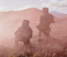 Two soldiers, both in Army uniform and kneeling at different heights are surrounded by pink smoke in the field. They are surrounded by dry grass and rolling hills in the background.