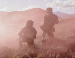 Two soldiers, both in Army uniform and kneeling at different heights are surrounded by pink smoke in the field. They are surrounded by dry grass and rolling hills in the background.