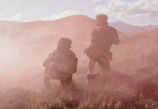Two soldiers, both in Army uniform and kneeling at different heights are surrounded by pink smoke in the field. They are surrounded by dry grass and rolling hills in the background.