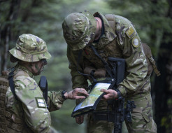Two uniformed aviators look at a map in the bush. The image has a very green feel - green uniforms and green surrounding bush. One aviator is looking down and you can't see their face. The other, only the side of their face (they have camo on). The image 