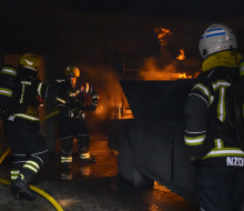 Personnel in firefighting uniform fight fire in a dark area. There is a simulated car fire. One person has his back to the camera and then there are two others to the left of the image.