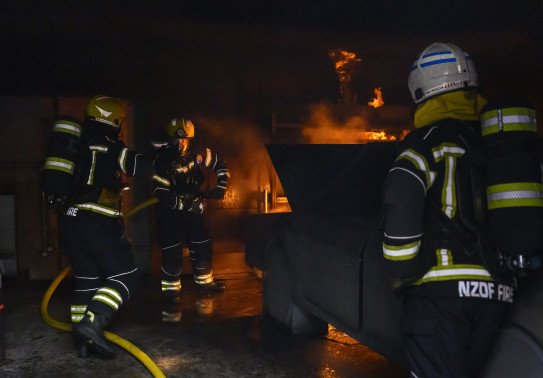 Personnel in firefighting uniform fight fire in a dark area. There is a simulated car fire. One person has his back to the camera and then there are two others to the left of the image.
