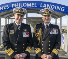Two Navy Chiefs stand side by side in front of a wharf that have an archway that reads, "HMNZS PHILOMEL LANDING".