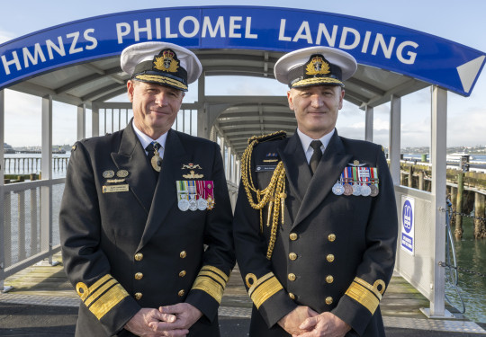 Two Navy Chiefs stand side by side in front of a wharf that have an archway that reads, "HMNZS PHILOMEL LANDING".