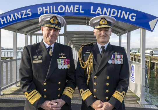 Two Navy Chiefs stand side by side in front of a wharf that have an archway that reads, "HMNZS PHILOMEL LANDING".