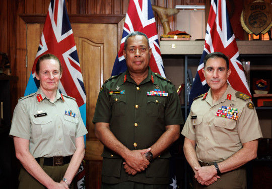 Image of three uniformed personnel standing in a line, in front of flags. All are looking at the camera smiling slightly with their hand together in front of them. 
