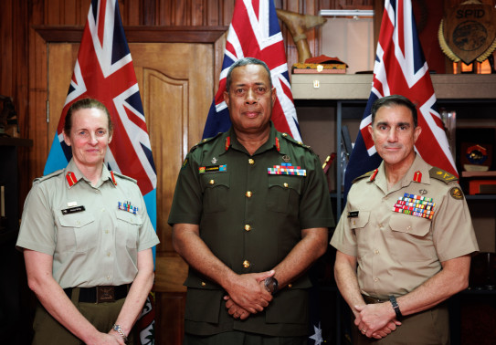 Image of three uniformed personnel standing in a line, in front of flags. All are looking at the camera smiling slightly with their hand together in front of them. 