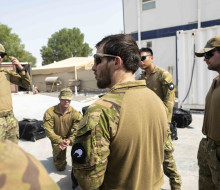 Soldiers stand in a circle for a brief, one of the soldiers is knelt down, containers, tents a prefabricated buildings surround the team under harsh sunlight.