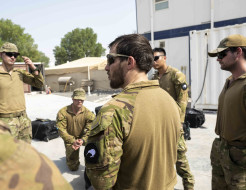 Soldiers stand in a circle for a brief, one of the soldiers is knelt down, containers, tents a prefabricated buildings surround the team under harsh sunlight.