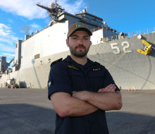 LT Cunningham stands with arms crossed in front of the large grey US Navy ship, USS Pearl Harbor. The number 52 is painted on the front-side of the ship.
