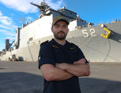 LT Cunningham stands with arms crossed in front of the large grey US Navy ship, USS Pearl Harbor. The number 52 is painted on the front-side of the ship.