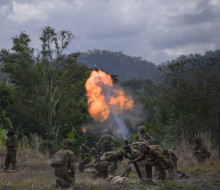 Army crews fire a mortar in a field in front of trees. The soldiers firing, are crouched down as a large flame and a round is launched from the mortar.