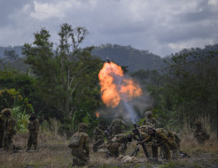 Army crews fire a mortar in a field in front of trees. The soldiers firing, are crouched down as a large flame and a round is launched from the mortar.
