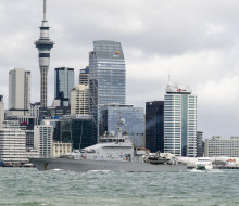 A grey ship sails in front of the Auckland city skyline.