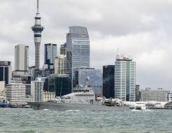 A grey ship sails in front of the Auckland city skyline.
