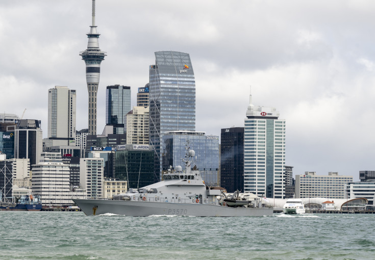 A grey ship sails in front of the Auckland city skyline.