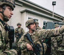 Soldiers are in between vehicles out the front of a white panelled single-story building, one soldier points to the right while others listen.