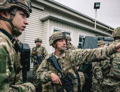 Soldiers are in between vehicles out the front of a white panelled single-story building, one soldier points to the right while others listen.