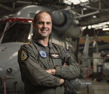 Male wearing an aircrew uniform smiles as he stands with his arms folded in front of a grey military helicopter. 