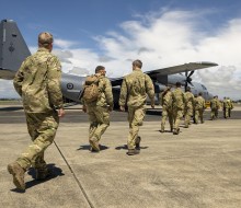 A line of personnel in military uniform, some with backpacks, walk across the tarmac at an airport towards a large grey military aircraft as they prepare to be deployed.