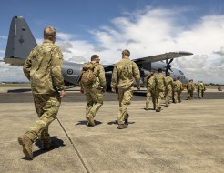 A line of personnel in military uniform, some with backpacks, walk across the tarmac at an airport towards a large grey military aircraft as they prepare to be deployed.