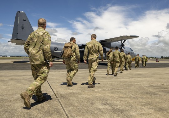 A line of personnel in military uniform, some with backpacks, walk across the tarmac at an airport towards a large grey military aircraft as they prepare to be deployed.