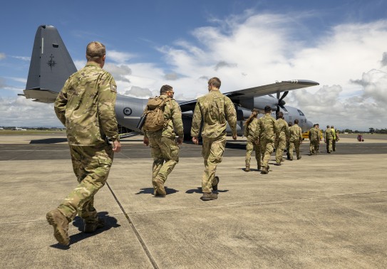 A line of personnel in military uniform, some with backpacks, walk across the tarmac at an airport towards a large grey military aircraft as they prepare to be deployed.
