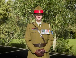 A male Army Officer stands smiling at the camera, he wears green ceremonial dress including leather gloves and full brimmed hat, his chest is adorned with medals
