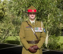 A male Army Officer stands smiling at the camera, he wears green ceremonial dress including leather gloves and full brimmed hat, his chest is adorned with medals