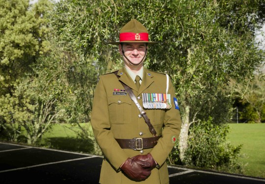 A male Army Officer stands smiling at the camera, he wears green ceremonial dress including leather gloves and full brimmed hat, his chest is adorned with medals