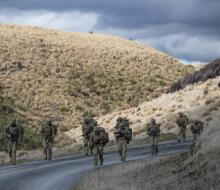 Soldiers conduct a pack march along a road that weaves between hills.