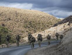 Soldiers conduct a pack march along a road that weaves between hills.