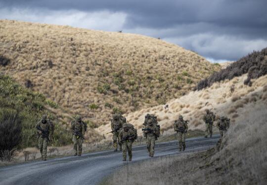 Soldiers conduct a pack march along a road that weaves between hills.