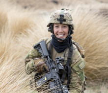 A woman smiles as she stands in long brown grass holding a black rifle and full camouflage army uniform including a helmet.