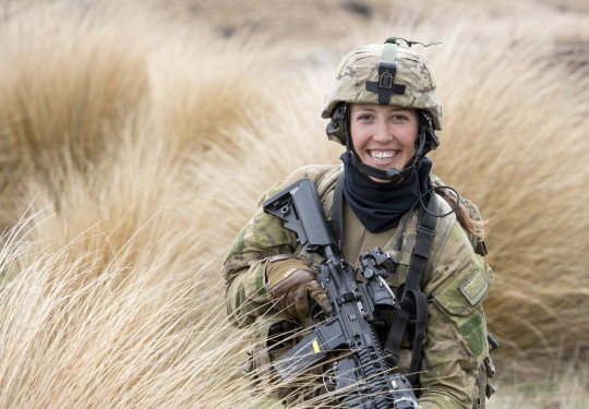 A woman smiles as she stands in long brown grass holding a black rifle and full camouflage army uniform including a helmet.