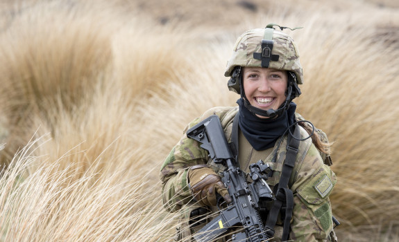 A woman smiles as she stands in long brown grass holding a black rifle and full camouflage army uniform including a helmet.