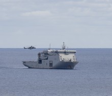 A large grey naval ship moves through the waters as a helicopter hovers above.
