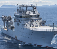 Stock photo of HMNZS Manawanui gliding through the ocean on a sunny day with land and hills in the distance. The ship is grey and rather bulky with a large crane on the back.