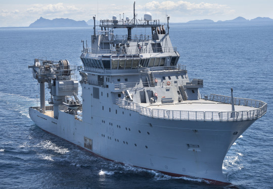 Stock photo of HMNZS Manawanui gliding through the ocean on a sunny day with land and hills in the distance. The ship is grey and rather bulky with a large crane on the back.