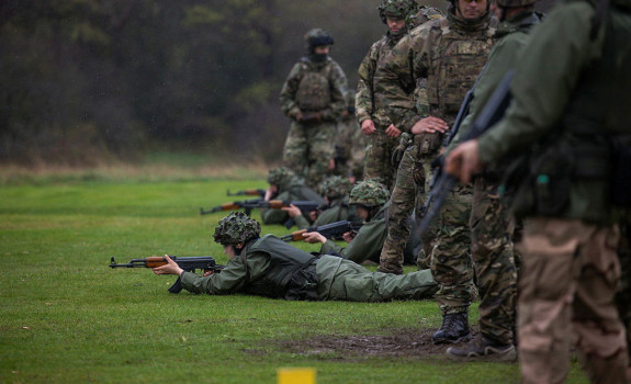 Soldiers in green and camouflage uniforms fire rifles down the range as more soldier stand behind them. 