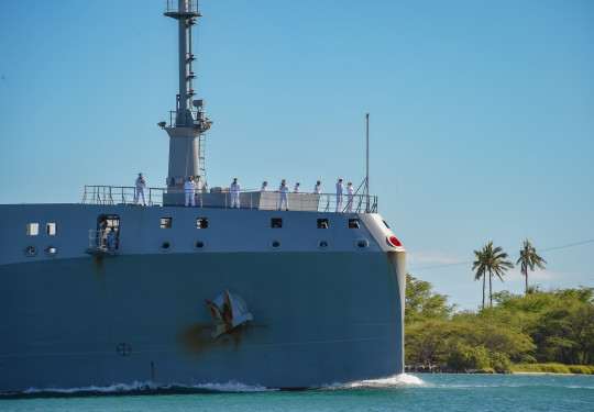 HMNZS Aotearoa sailing into Hawaii's Pearl Harbour on a sunny day with palm trees featuring in the background.