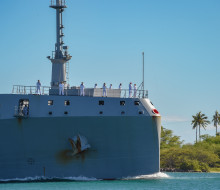 HMNZS Aotearoa sailing into Hawaii's Pearl Harbour on a sunny day with palm trees featuring in the background.