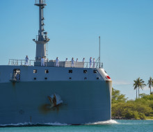 HMNZS Aotearoa sailing into Hawaii's Pearl Harbour on a sunny day with palm trees featuring in the background.