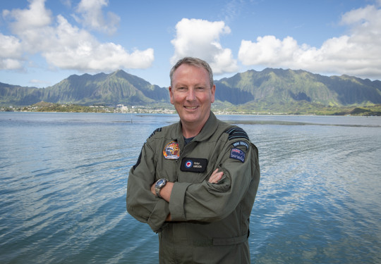 Group Captain Pete Gibson in Hawaii, stands with arms crossed and a smile in front of a body of water and hills in the background. 